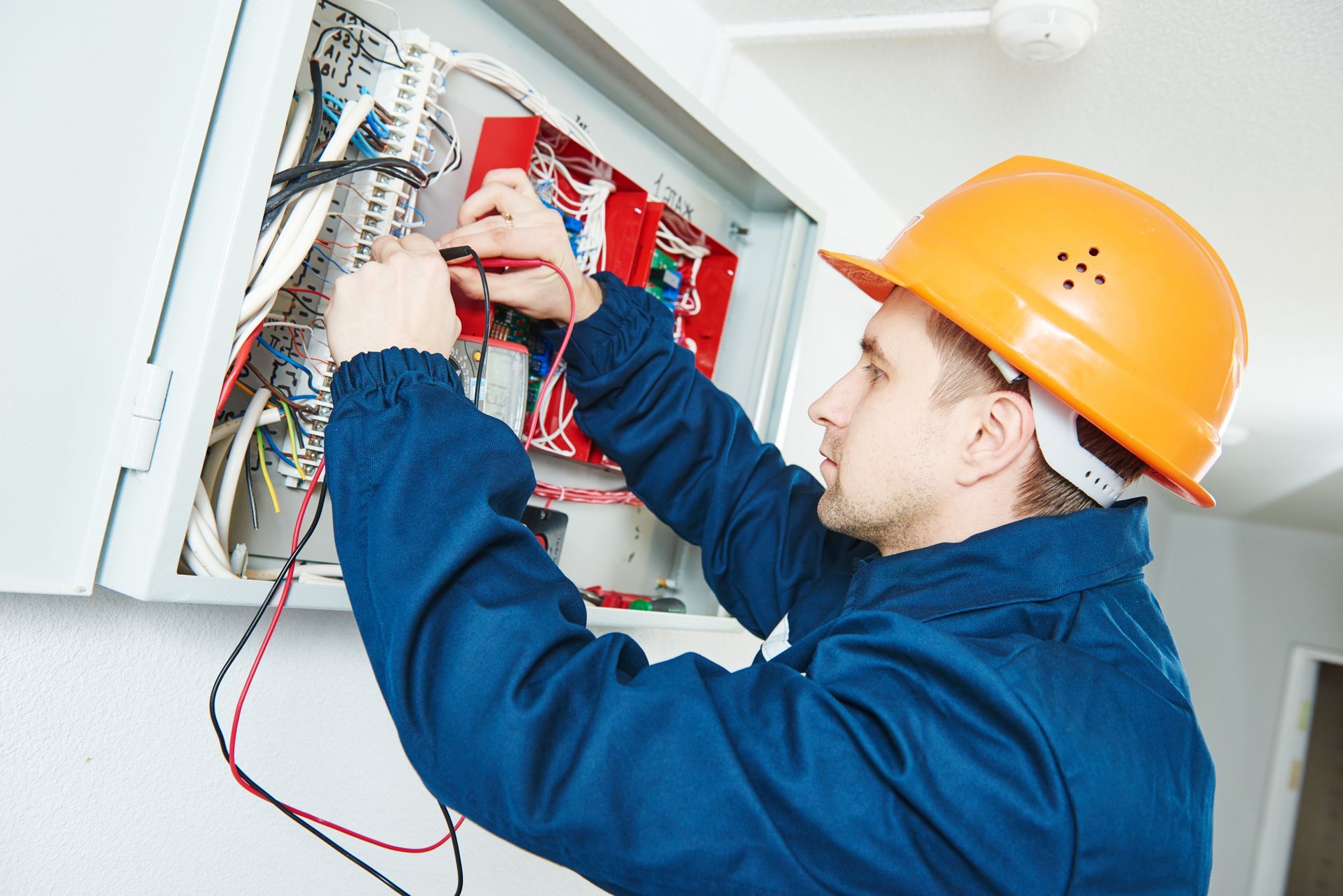 An electrician in a blue uniform and orange hard hat uses a multimeter to test wiring inside an electrical panel.