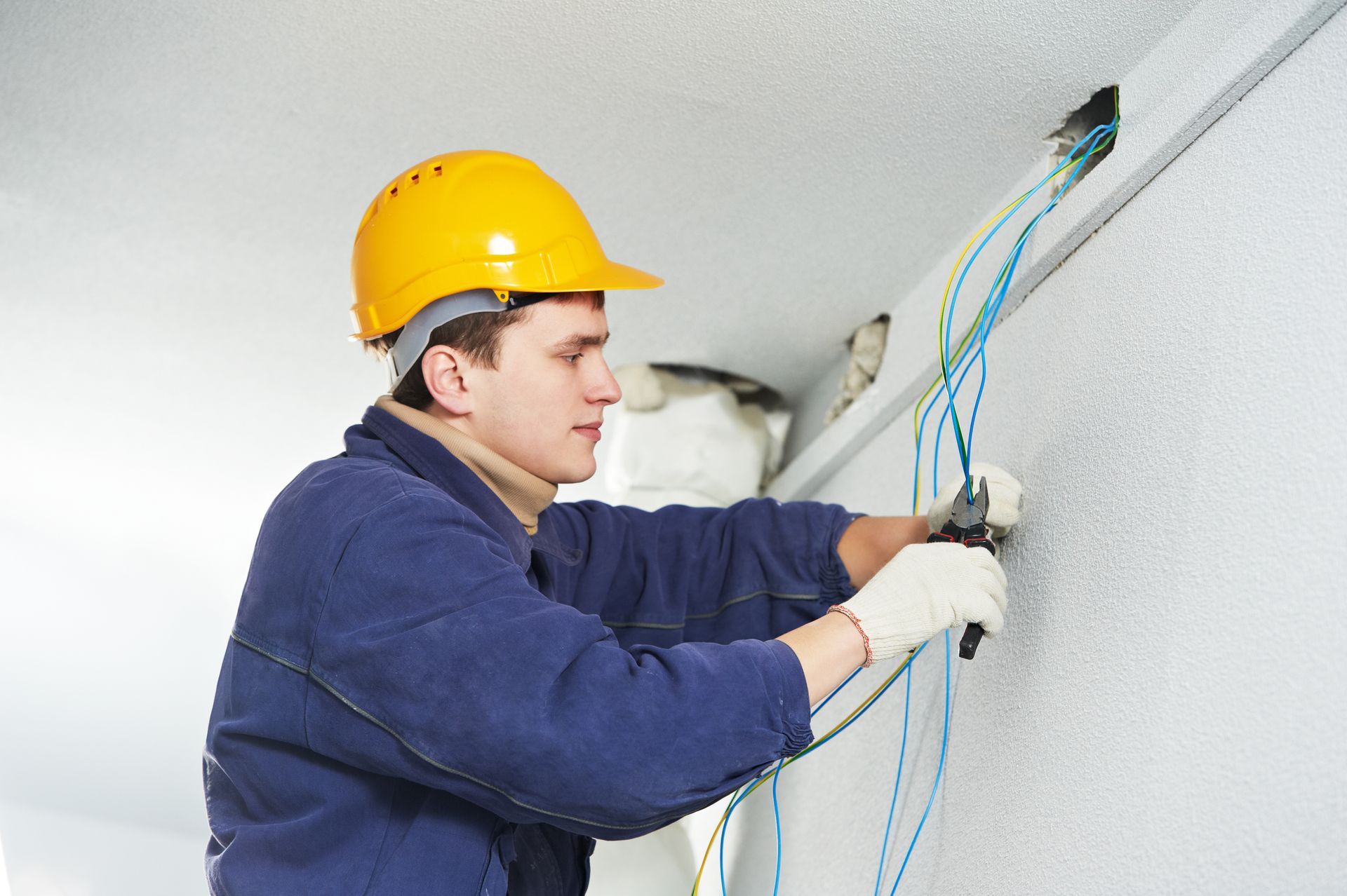 An electrician in a yellow hard hat and blue uniform uses pliers to work on electrical wires protruding from a wall.