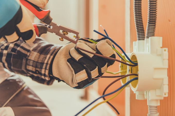 An electrician wearing work gloves uses wire strippers on electrical wires connected to a wall-mounted box.