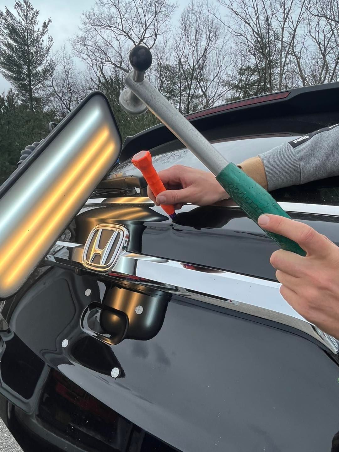 Hands using tools to remove a dent from a black Honda car trunk.
