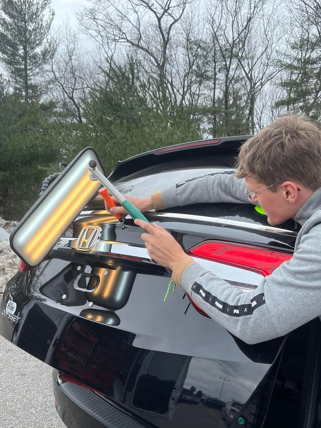 A person uses a dent puller tool on a black car's rear, outdoors.