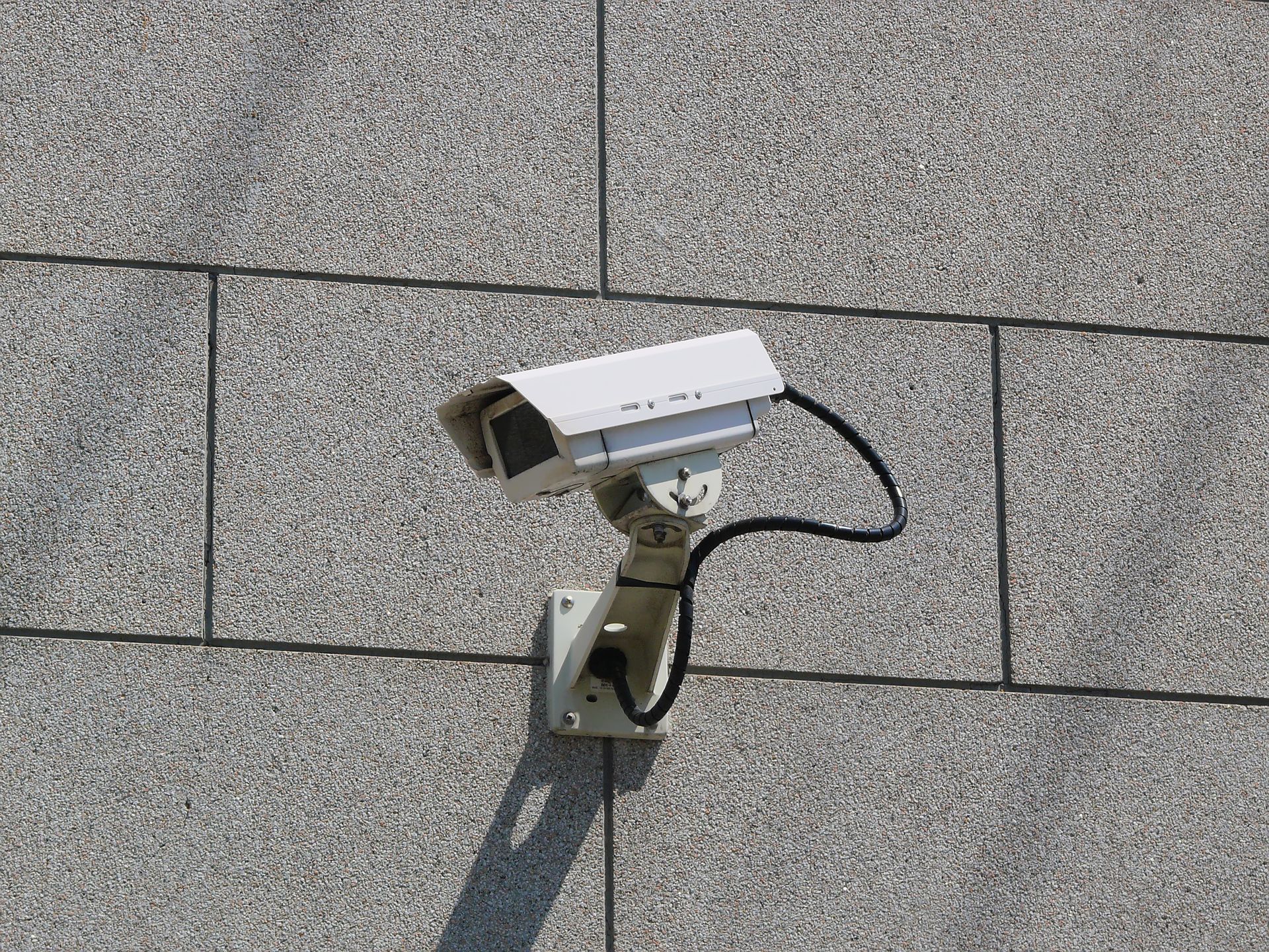 Security camera mounted on a gray, stone-tiled building, aimed downwards.