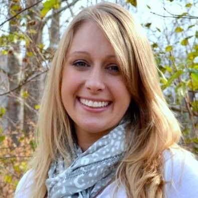 Blonde woman smiling with light blue scarf, trees in background.