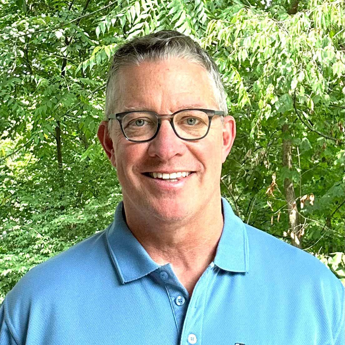 Man with glasses smiling, wearing a blue polo shirt, in front of green foliage.