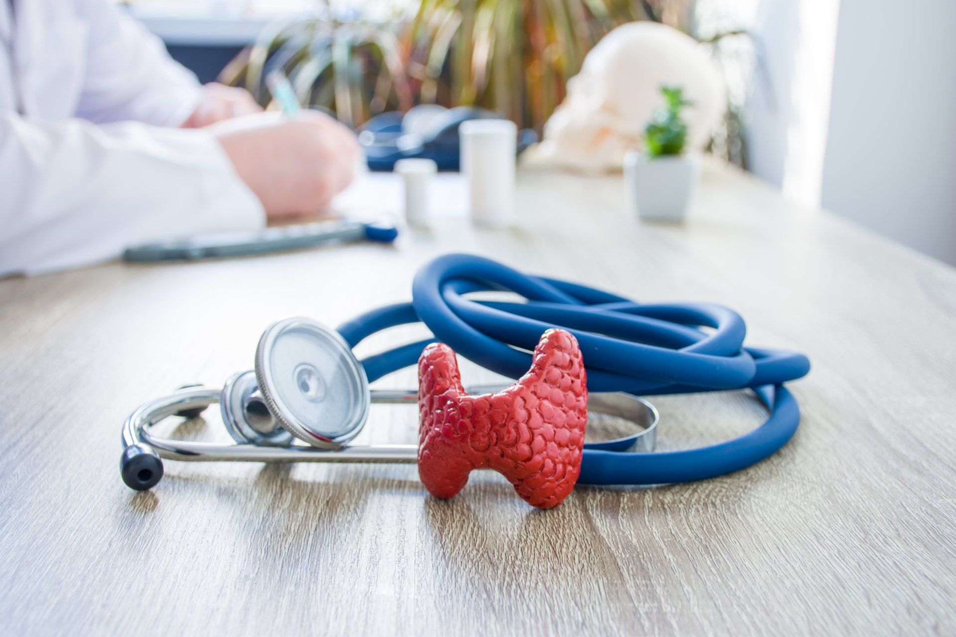 Red thyroid model and blue stethoscope on a wooden desk with a doctor writing in the background.