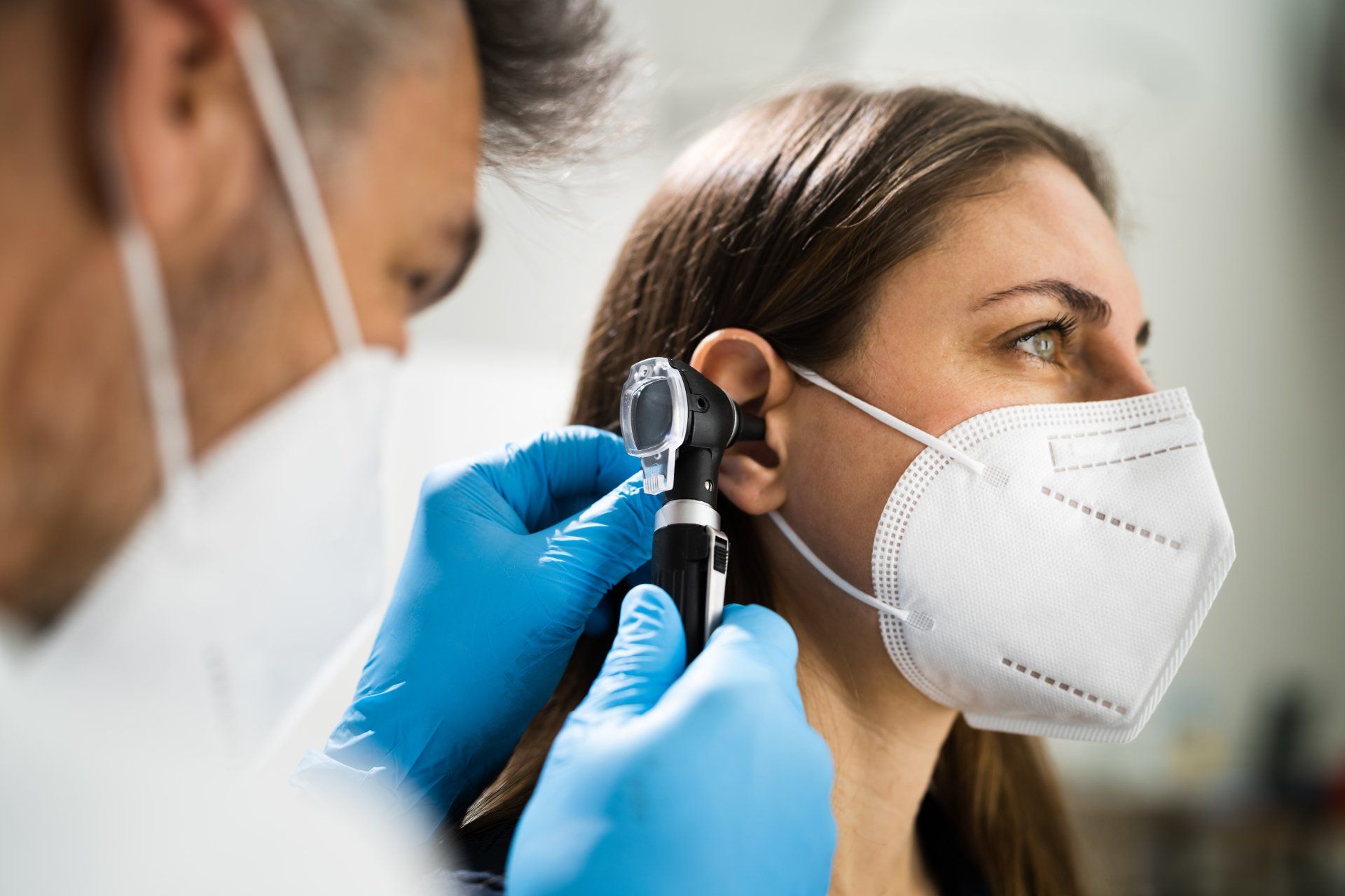 Doctor examining a patient's ear with an otoscope. Both wear masks and gloves.