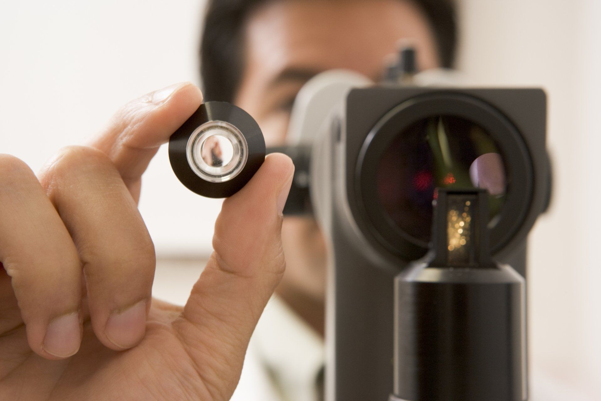 Person holding an eye lens, with an eye exam machine in the background, indoor setting.