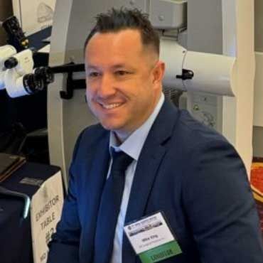 Man in a suit smiling; sitting at an exhibitor table with medical equipment in the background.