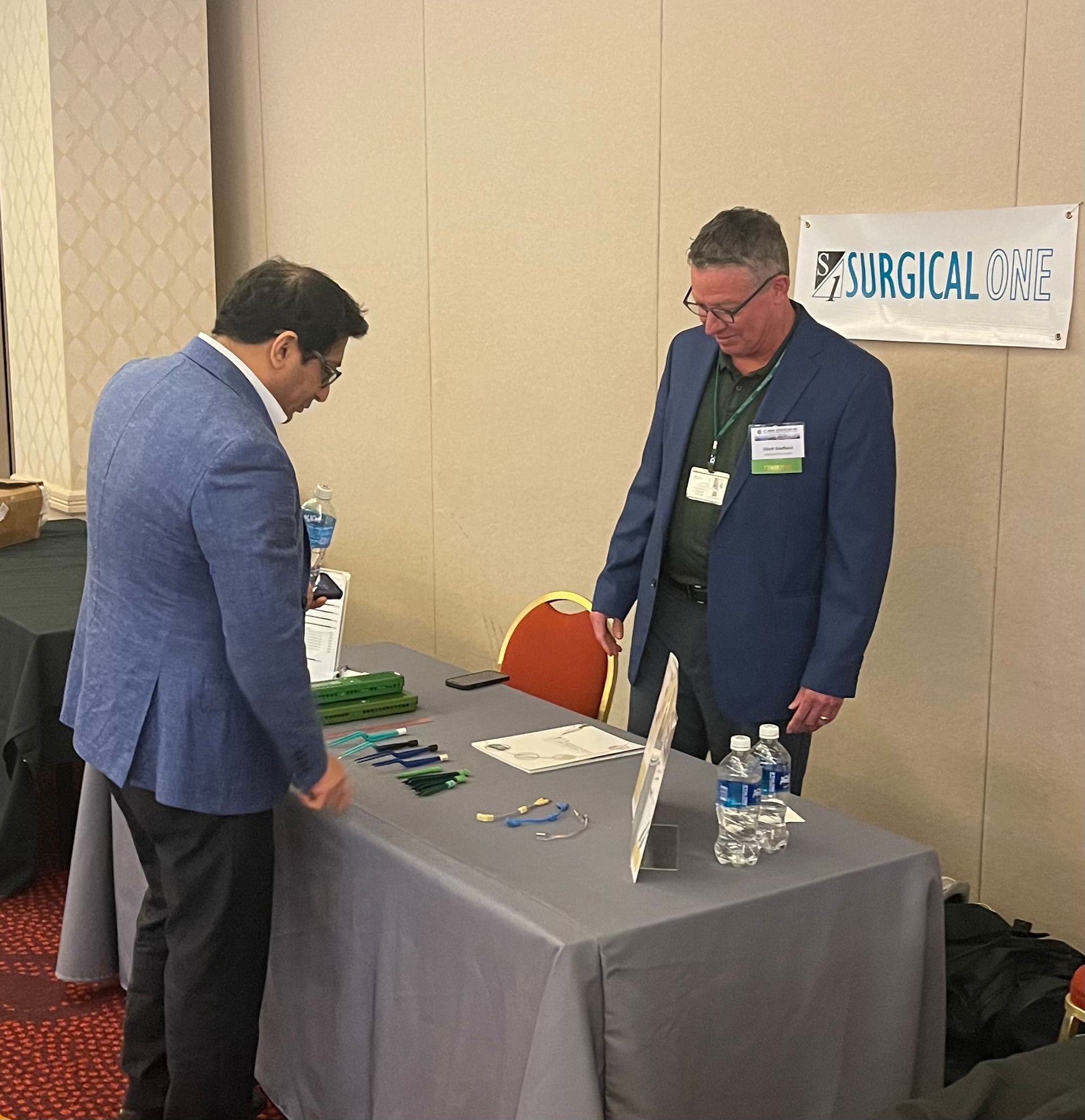 Two men at a trade show table, examining surgical tools. Sign for Surgical One.