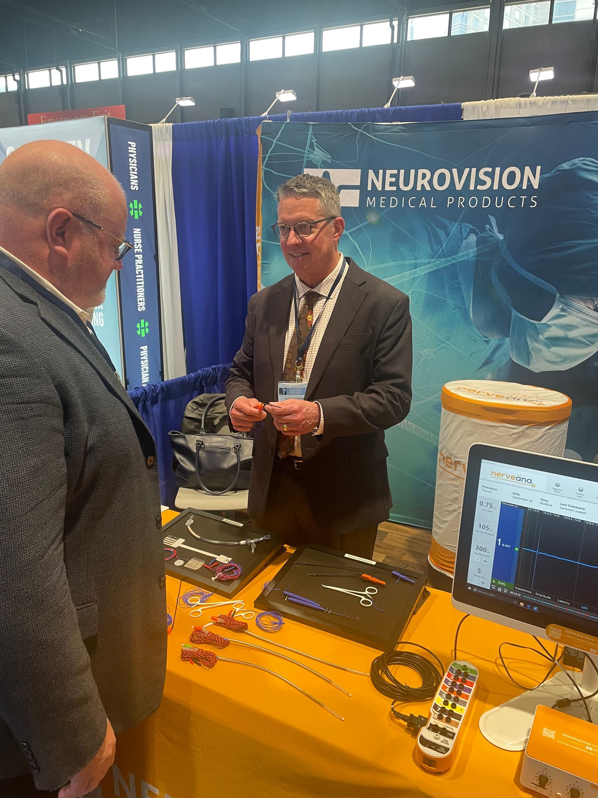 Man at a trade show booth for Neurovision, showing a product to a visitor. Displays and a computer are on the table.