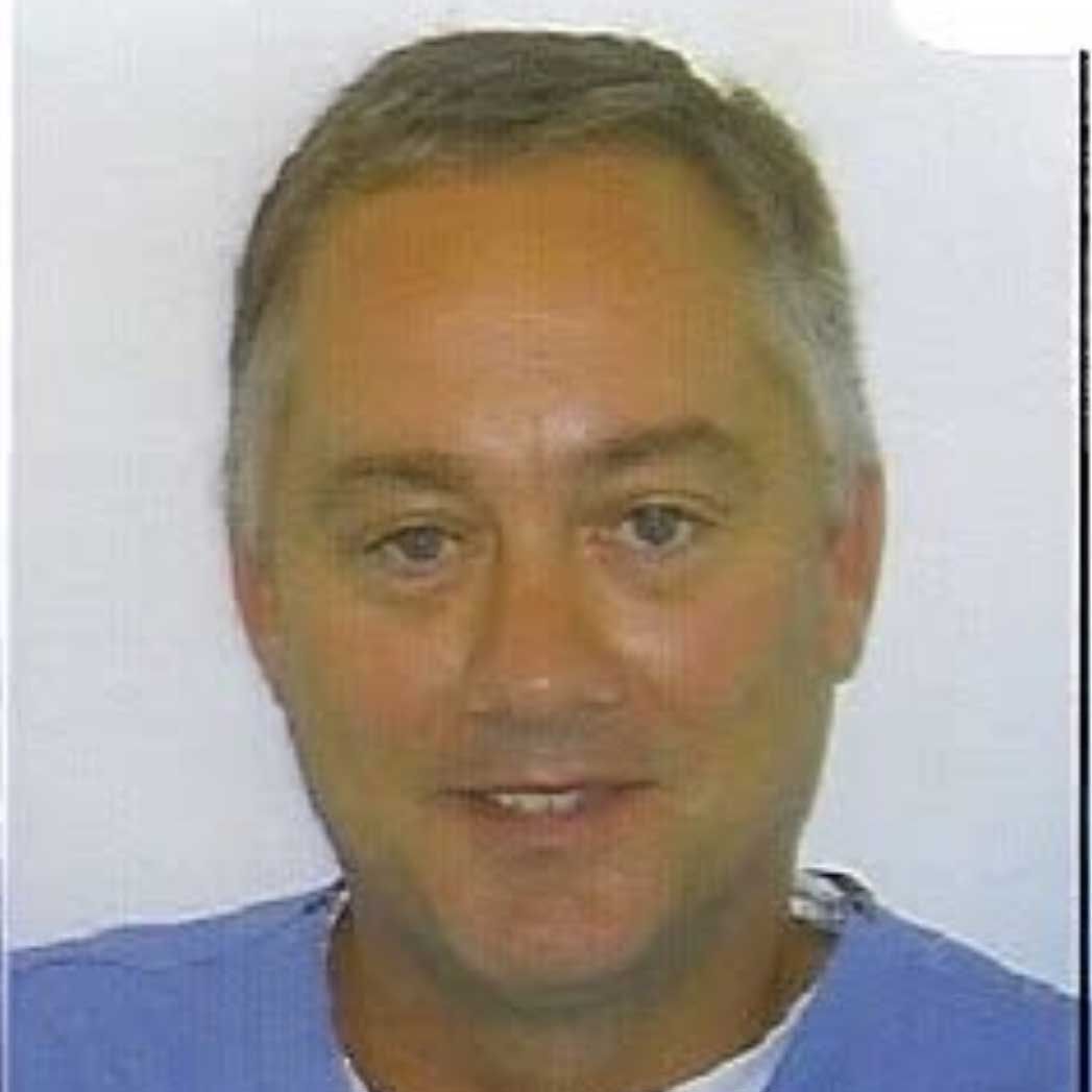 A fair-skinned man in a blue shirt smiles at the camera. Graying hair, eyes open, neutral setting.