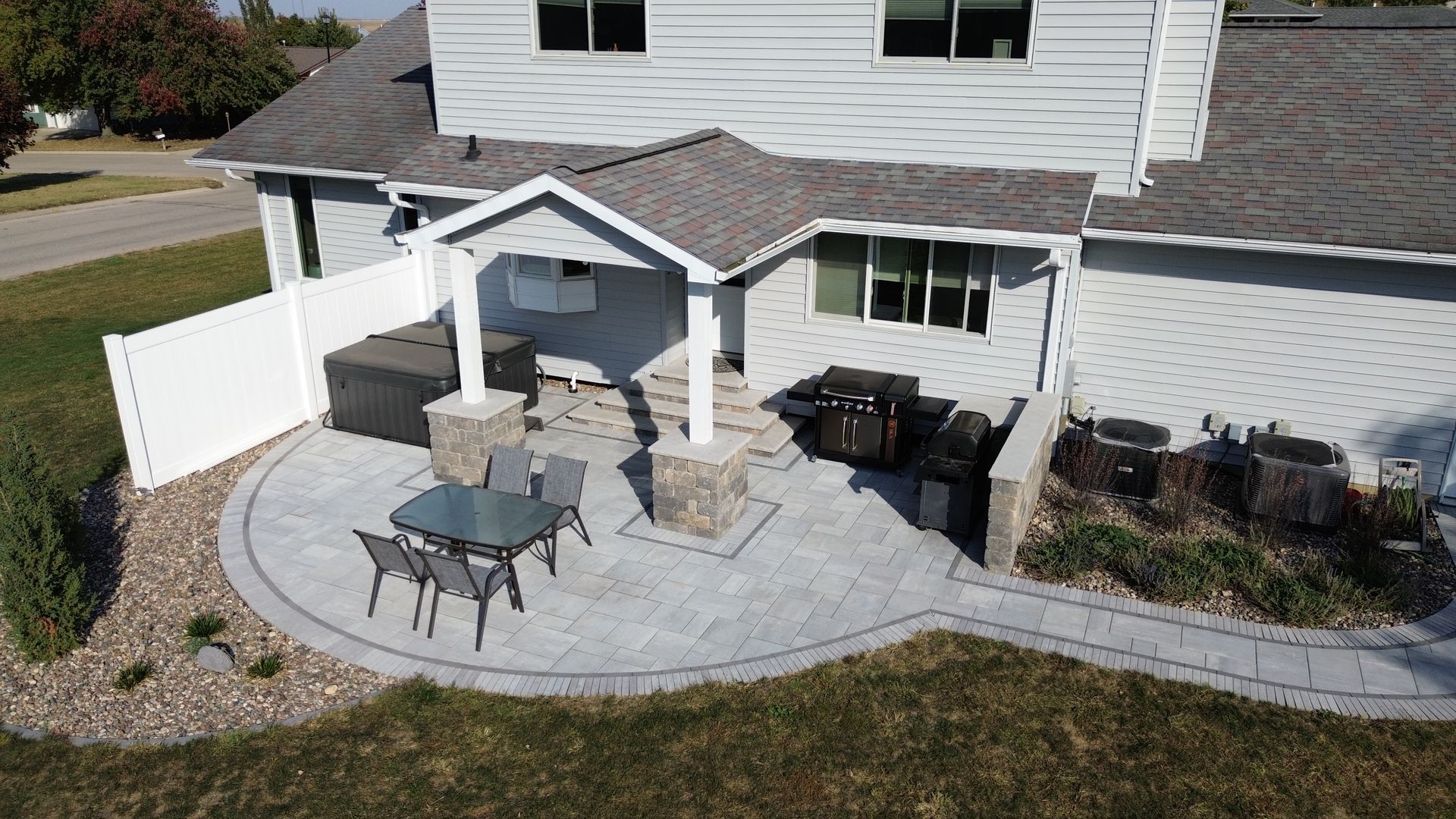 Backyard patio with gray pavers, built-in grill, seating area, and a white fence.