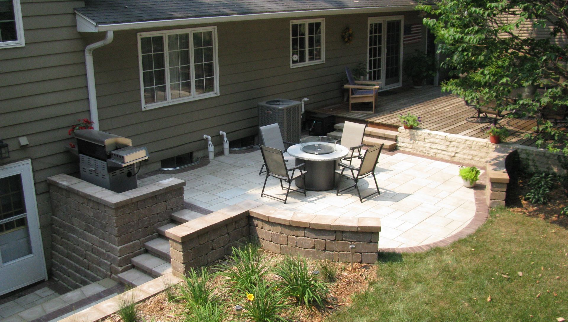 Backyard patio with fire pit, seating, and built-in grill, adjacent to a house with a door and windows.