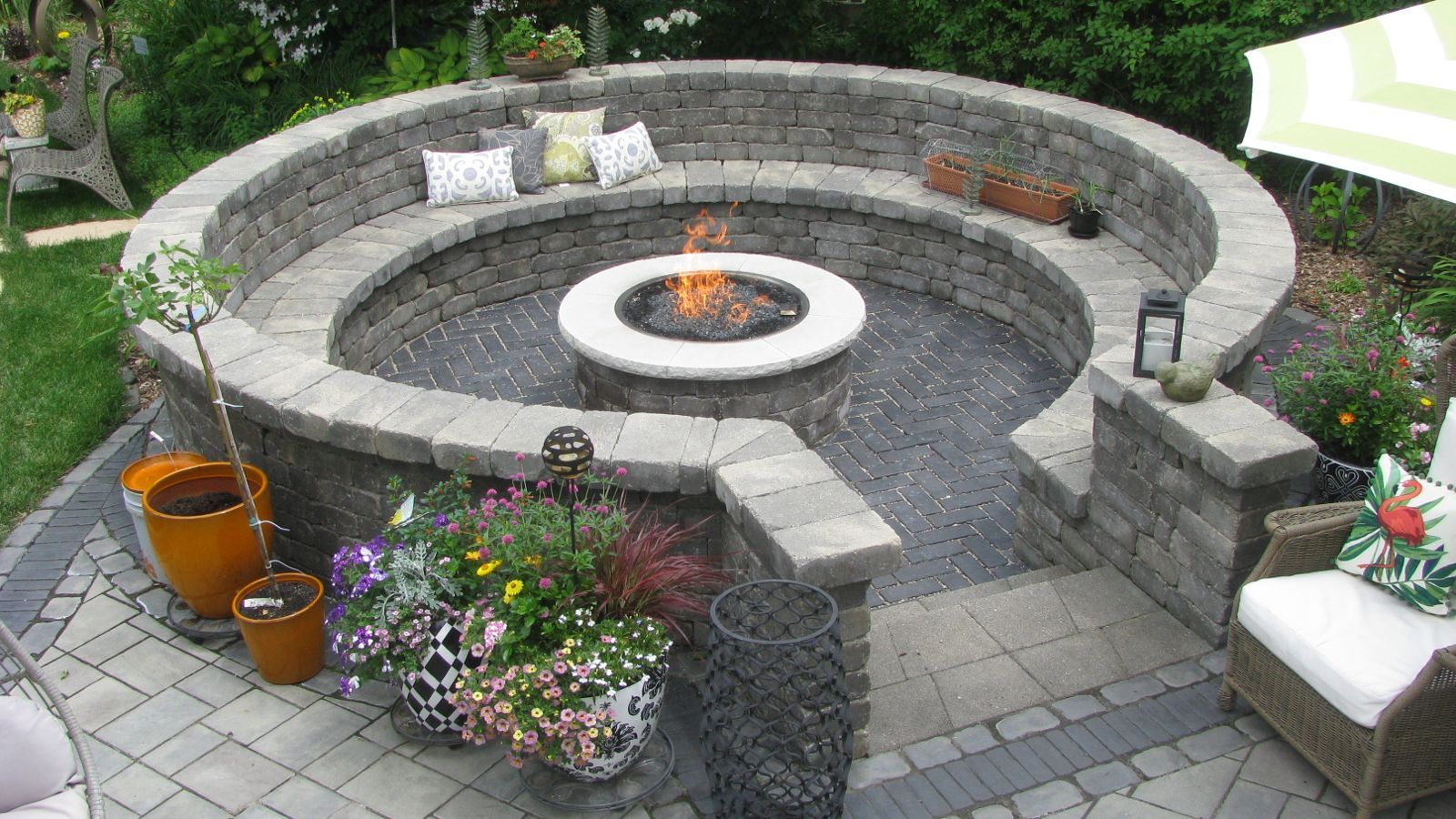 Circular stone fire pit with built-in seating on a patio, surrounded by plants, with a lit fire.