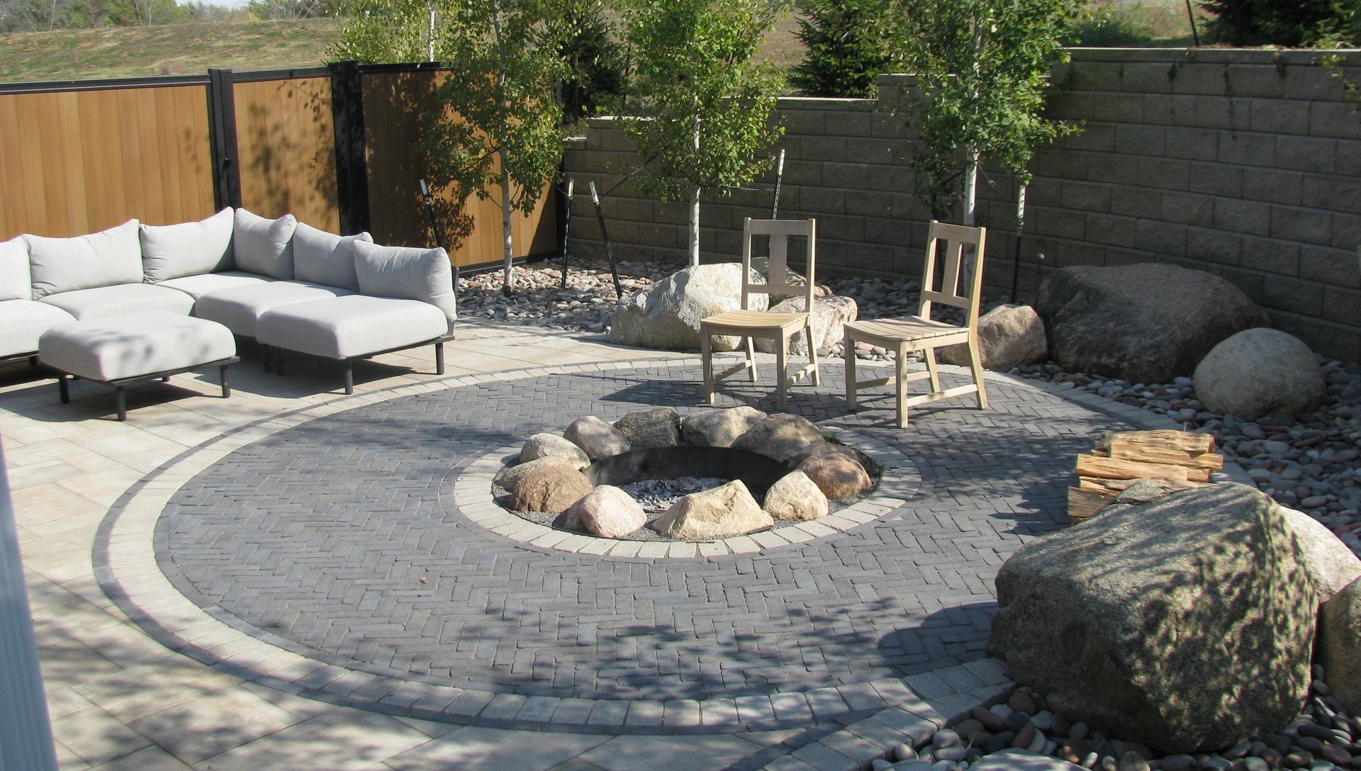 Outdoor patio with fire pit, seating area with sofa and chairs, surrounded by stones and trees.
