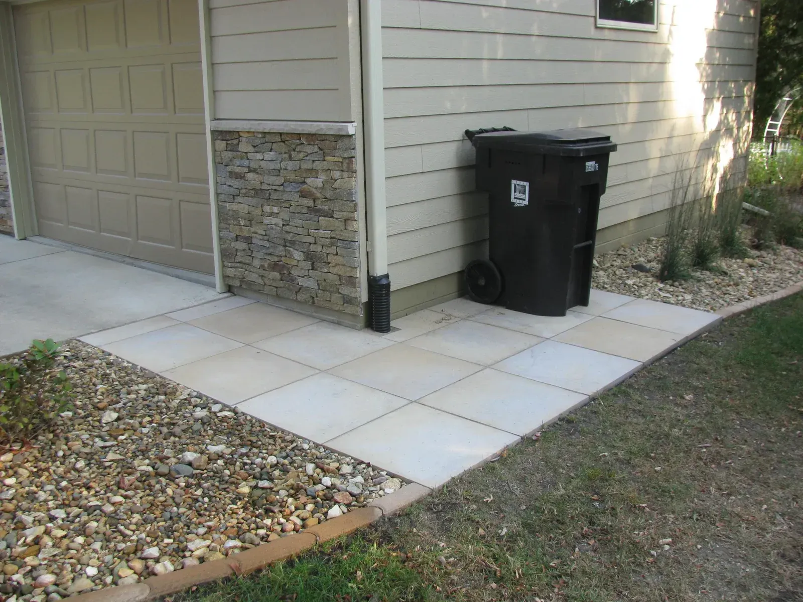 A black trash can sits on a sidewalk in front of a garage — Waterloo, IA — Matthias Landscaping Co