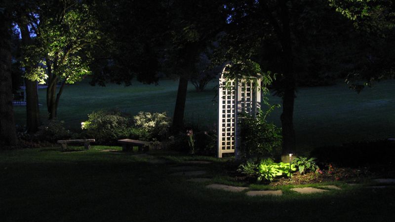 Nighttime garden scene with illuminated trees, benches, and a white trellis.