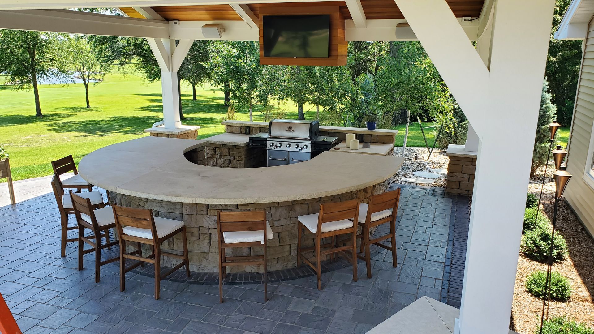 Outdoor kitchen with curved bar, grill, and TV. Surrounded by patio and green lawn.