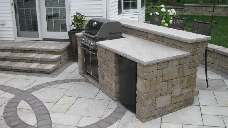 Outdoor kitchen with built-in grill, refrigerator, and countertop, constructed from beige stone, on a paved patio.