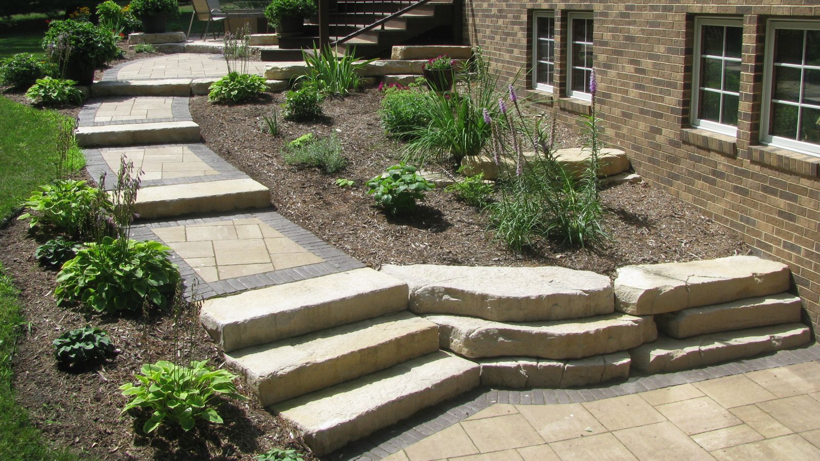 Stone steps and walkway leading up a tiered garden with various plants next to a brick building.