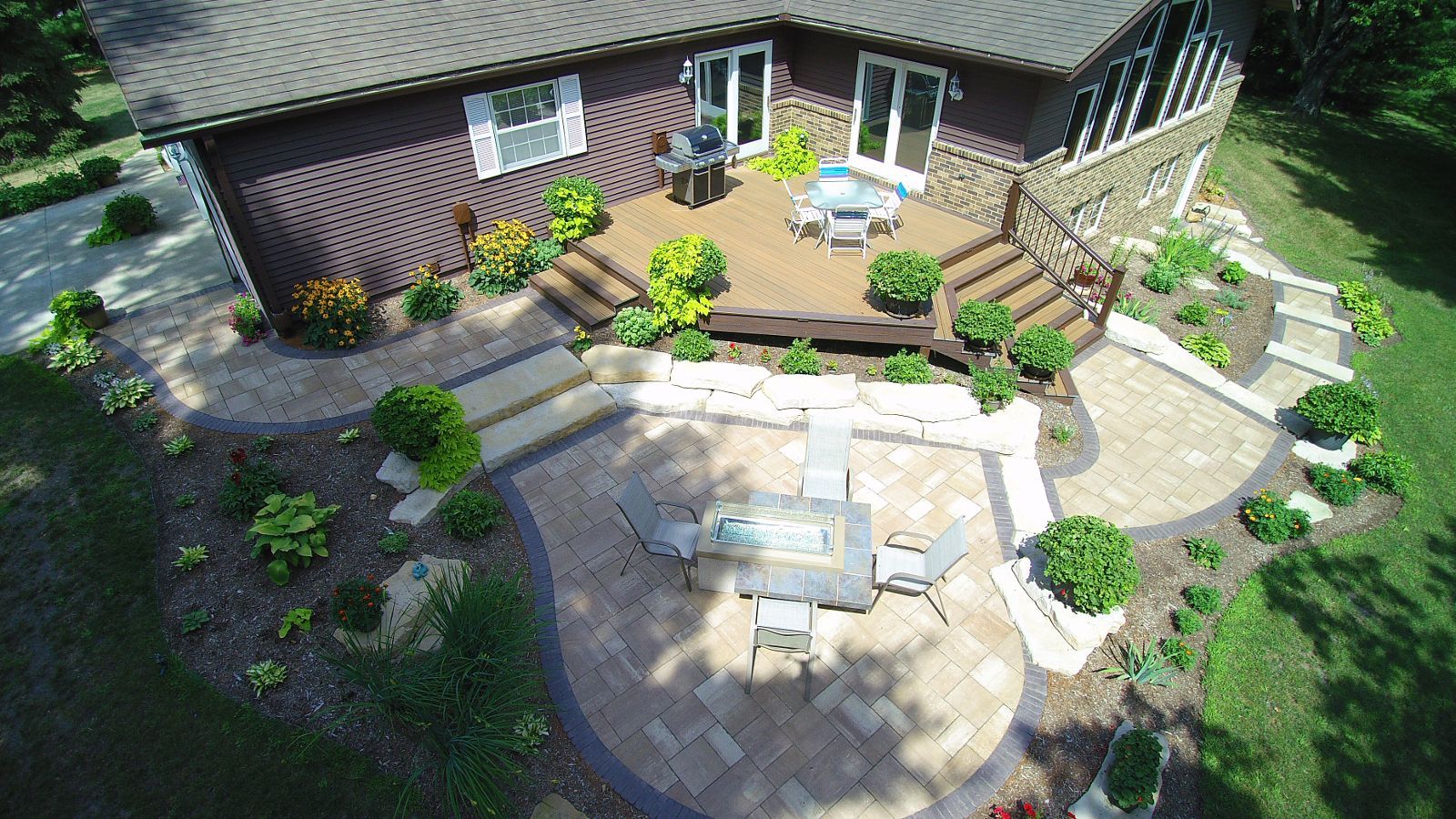 Aerial view of a home with a deck and patio. Brown house, dark wood deck, and paved patio with outdoor furniture.