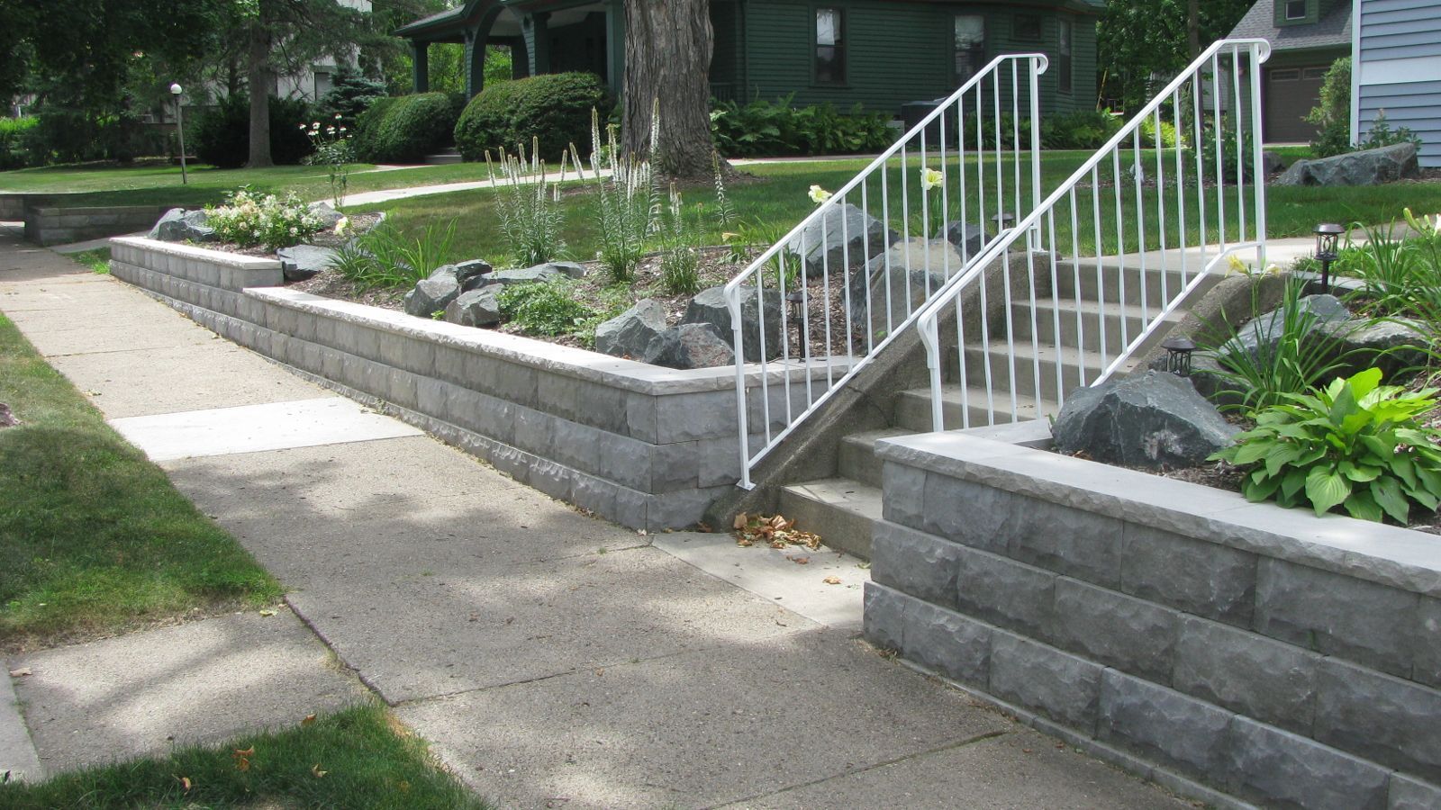 Concrete steps with white railing flanked by retaining walls with landscaping.
