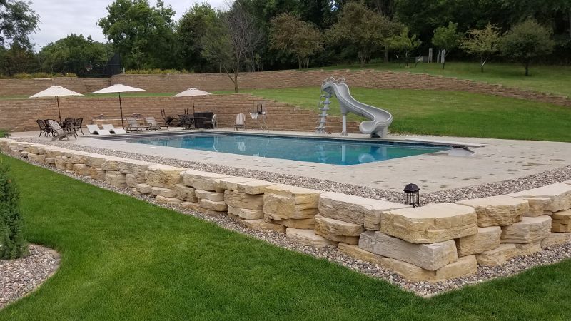 Rectangular swimming pool with slide and stone retaining wall. Lawn with umbrellas and chairs.