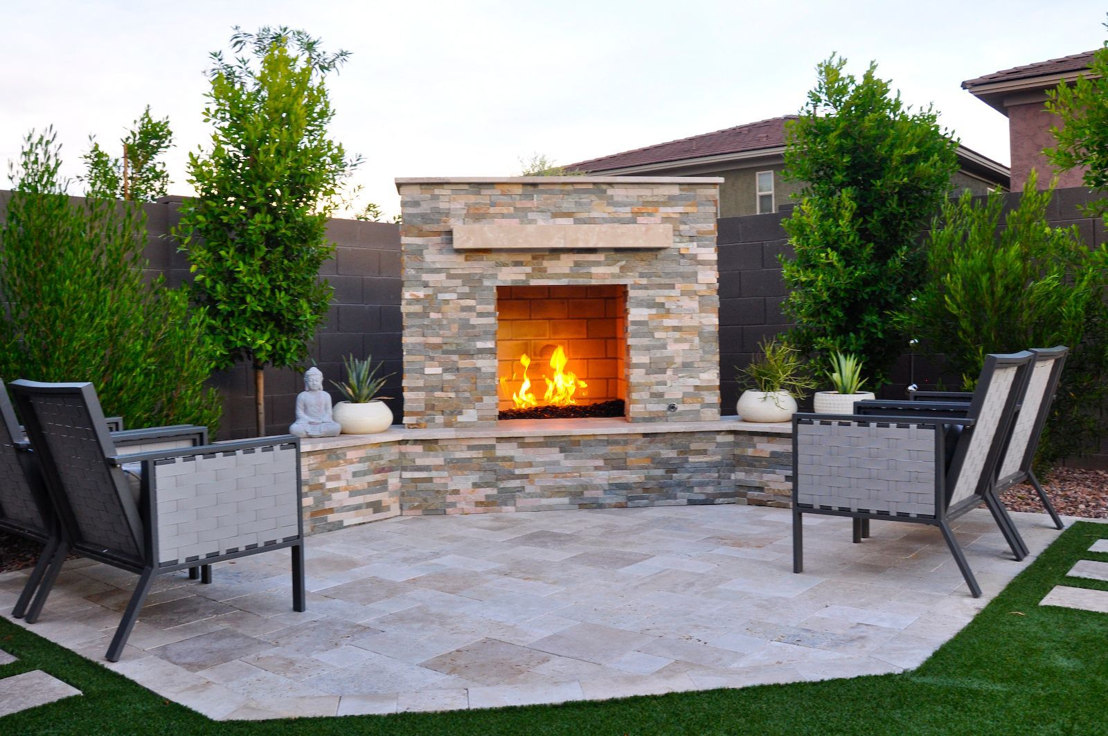 A stone fireplace is surrounded by chairs and trees in a backyard.