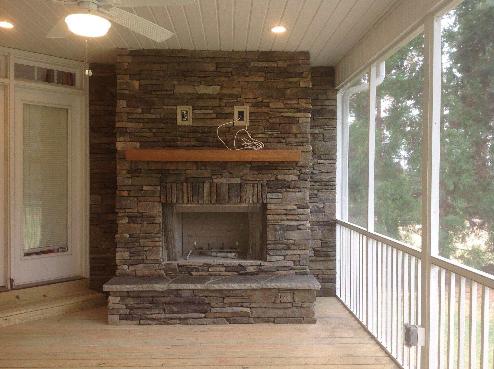 A screened in porch with a stone fireplace and a ceiling fan.
