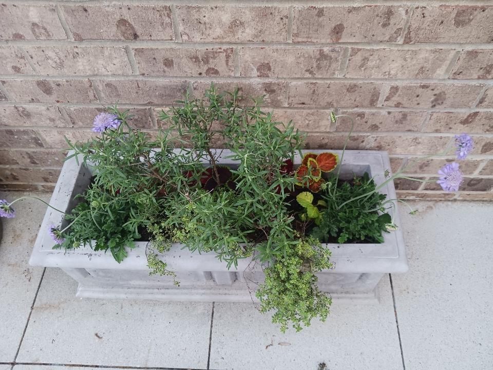 A planter filled with plants and flowers is sitting on a tiled floor in front of a brick wall.
