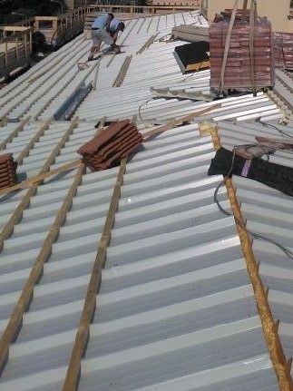 Una persona lavora su un tetto spiovente ricoperto di lamiere metalliche bianche e listelli di supporto in legno, con delle tegole impilate nelle vicinanze.