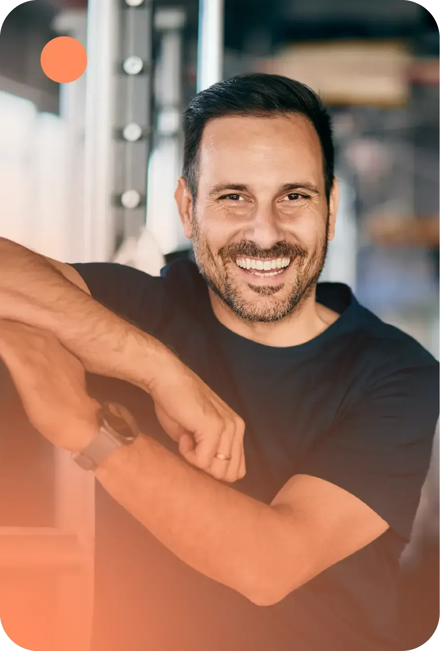 A smiling person with a beard, wearing a black t-shirt and a watch, leans against gym equipment.