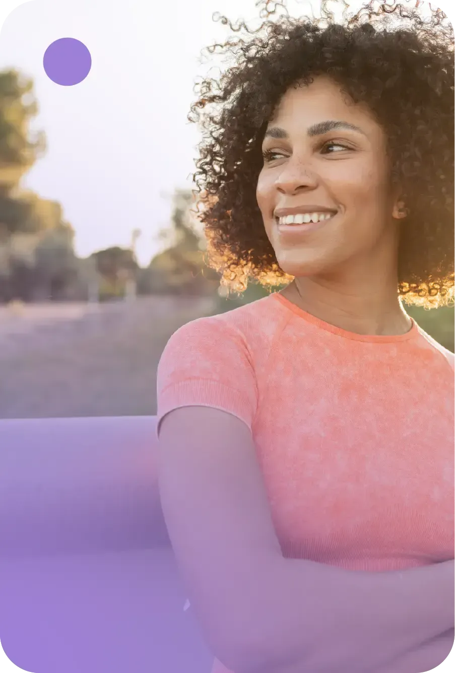 A smiling person with curly hair wears a coral shirt and carries a purple yoga mat in an outdoor setting at sunset.