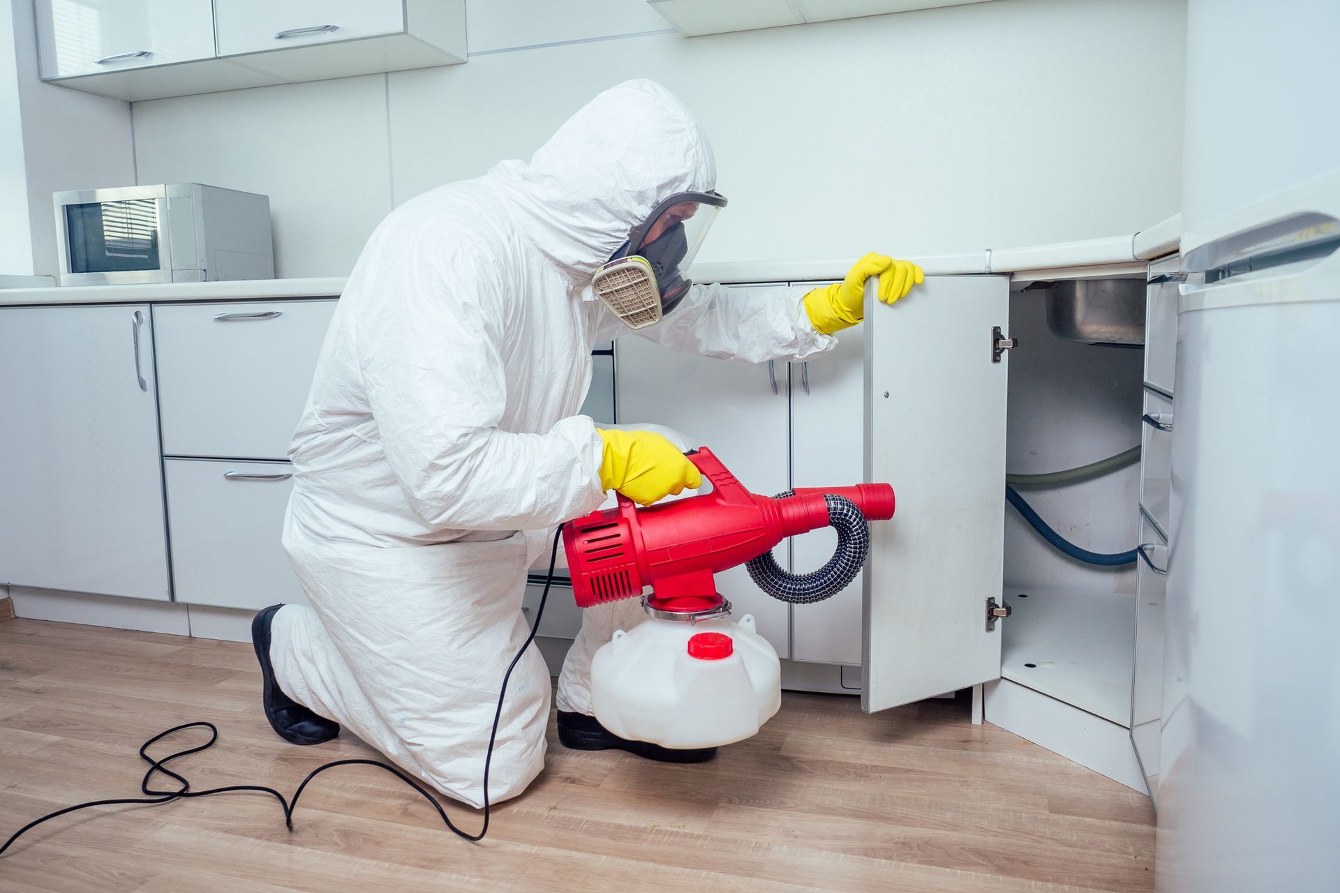 Pest exterminator in protective suit using fumigation machine inside residential kitchen.