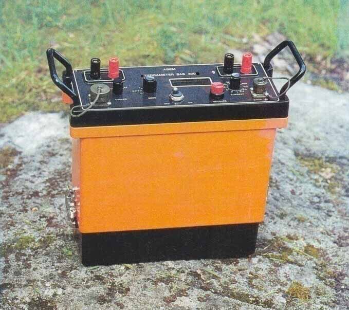 Dispositivo de prueba eléctrica de color naranja y negro, con asas, sobre una roca al aire libre.