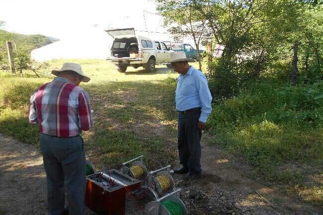 Dos hombres con sombreros examinan equipos en un camino de tierra, un camión blanco con la parte trasera abierta está detrás de ellos.