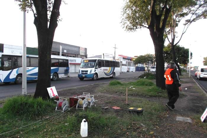 Autobuses en una calle con árboles y acera. Una persona con chaleco naranja camina junto a la carretera.