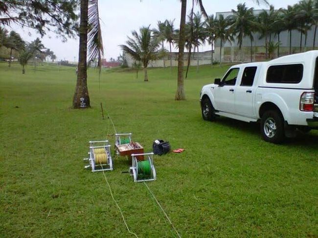 Un camión blanco estacionado en un campo de hierba con carretes de alambre y equipo. Árboles y edificios al fondo.