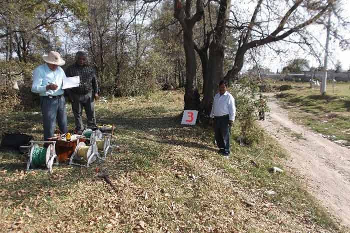 Tres personas inspeccionando un terreno; uno escribe en un portapapeles, uno está detrás y otro junto a un árbol con un cartel 