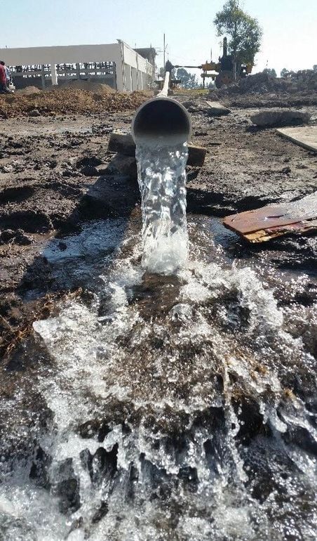 El agua brota de una tubería negra sobre un suelo fangoso; al fondo se ve una obra en construcción.