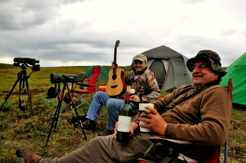 Two men are sitting in a field with a guitar and a bottle of wine