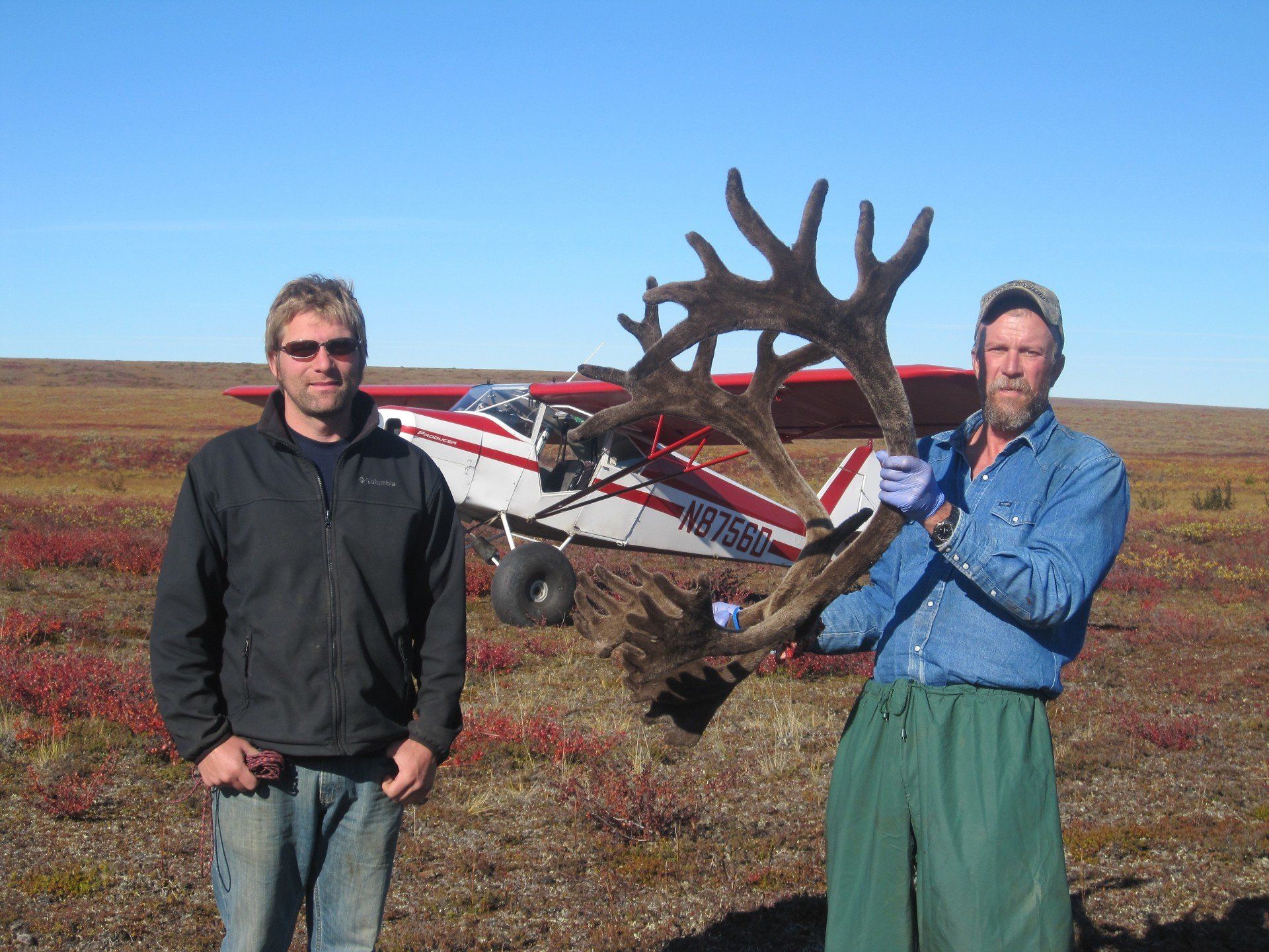 Two men holding a large antlers in front of a small plane