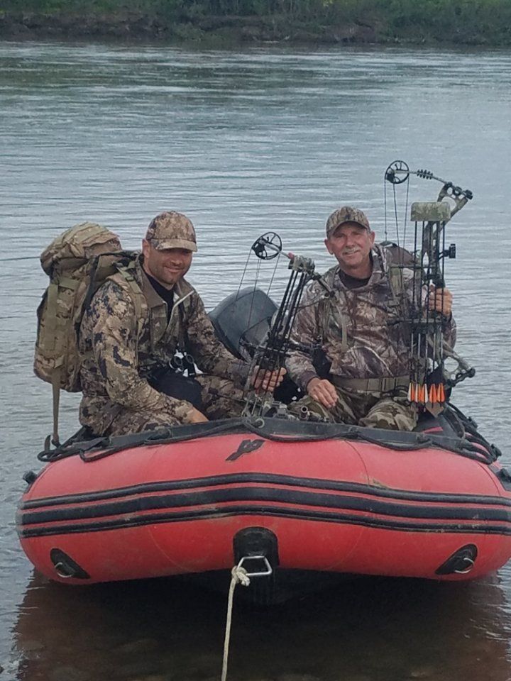 Two men are sitting in a boat in the water holding bows and arrows.