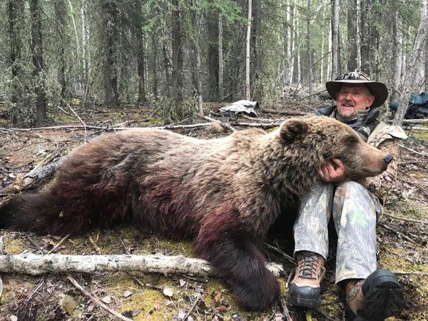 A man is sitting next to a large brown bear in the woods.