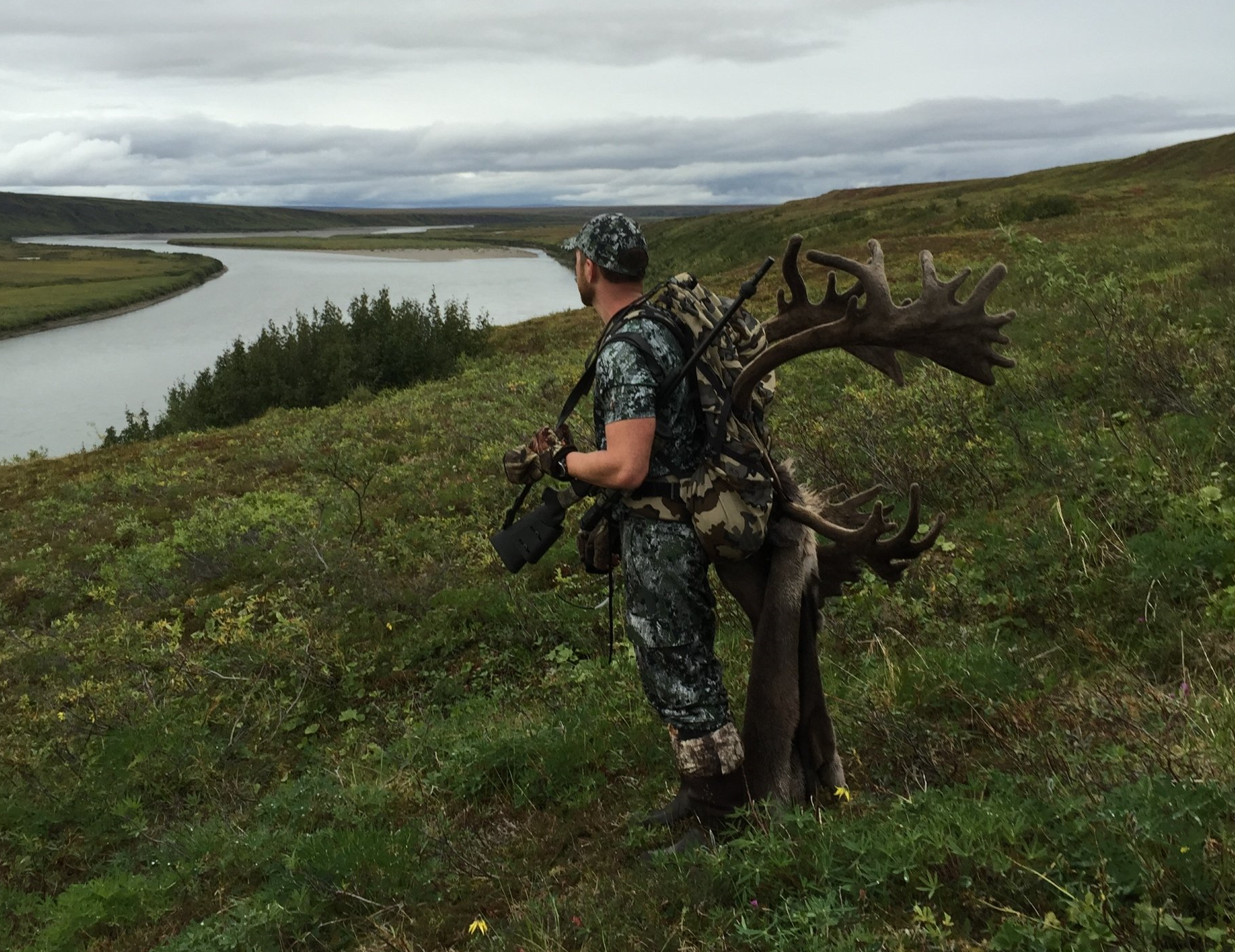 A man is standing next to a large deer in a field with a river in the background.