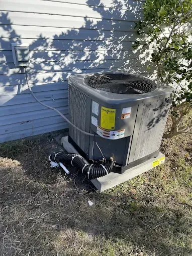Outdoor air conditioning unit next to a building, with connected black hoses and electrical box.