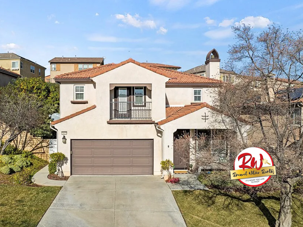 Two-story beige house with brown roof and garage, blue sky, and a real estate logo.