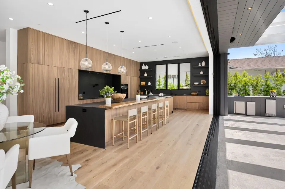 A contemporary home kitchen featuring wood cabinetry, a dark island with seating, and a wide opening to the outdoors.