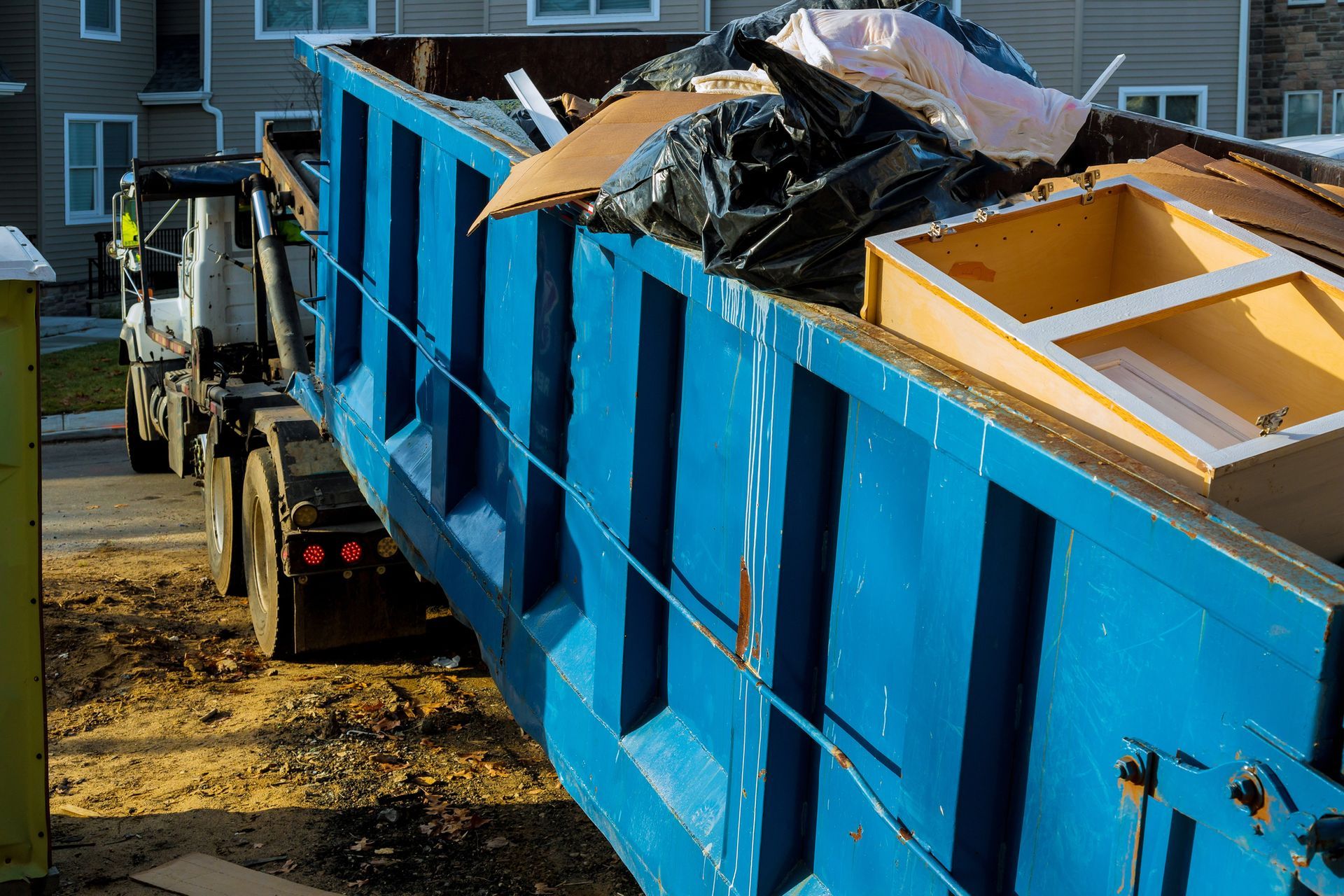 A large, blue construction dumpster filled with trash and a wooden cabinet, hitched to a truck outside a house.