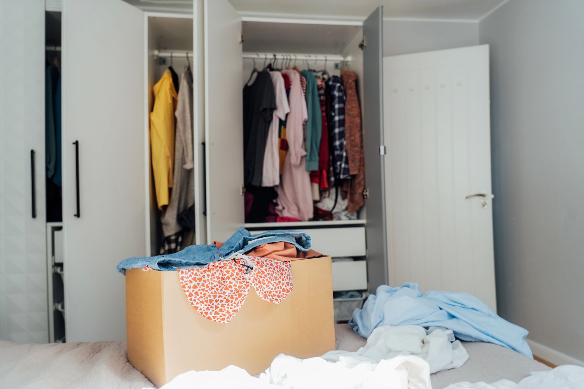 A box of clothes on a bed in a room with an open wardrobe filled with hanging garments.